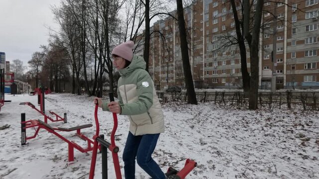 Young woman exercising on outdoor stepper in winter fitness park joyfully moving back and forth showing active healthy lifestyle outdoor cold weather training motion