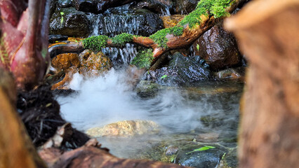 Artificial small waterfall and stream with rocks, moss, and tree branches. lush and green scene with misty effect at the bottom of the falls. decorative water garden features or natural settings. 