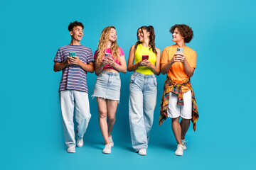 Fototapeta premium Funny group of four friends with colorful phones posing against a bright blue studio wall enjoying a casual fashion moment