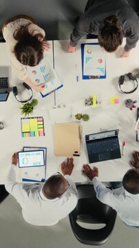Vertical Video Top down view zoom in of employees discussing business intel and performance in meeting. Multinational company work team reviews reports, forecasts and diagrams to align strategy