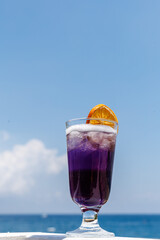 glass of purple drink with ice and orange slice on beachside railing. Blue sea and cloudy sky on background. Copy space, vertical