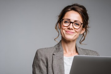 Stylish businesswoman wearing glasses smiling confidently with laptop against grey background
