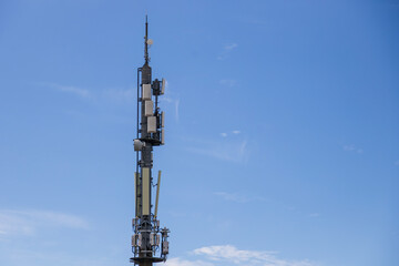 A tall cellular communication tower stands against a clear blue sky, equipped with numerous antennas and broadcasting equipment.