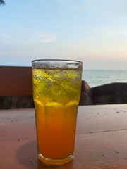 Close-up of a sweaty glass of cold orange or yellow iced drink on a wooden table with the blurry sea and sunset horizon in the background.
