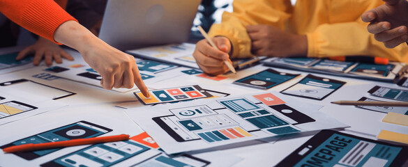A diverse team engages in a collaborative session, strategizing the design of a mobile application with colorful wireframes and various prototypes laid out on the table. SACTR