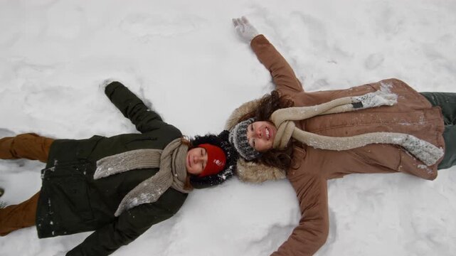 Top view portrait of happy young Caucasian mother and teenage son, wearing warm parka jackets and woolly hats falling on back in snow during winter walk, looking at camera and laughing