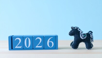 明るい室内のレトロな木の机の上に置かれた、2026年の数字が書かれた水色の木のブロックと、黒い羊毛フェルトの馬のぬいぐるみ。全体的に穏やかで希望に満ちた雰囲気。来年の干支