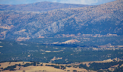 A vast and arid mountain valley landscape is seen from a high viewpoint showing the dry terrain and rolling hills under a blue sky