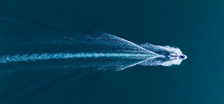Detailed top-down aerial view of a motorboat with people speeding across dark blue open water. The dynamic white wake design in the deep ocean creates striking geometric patterns.