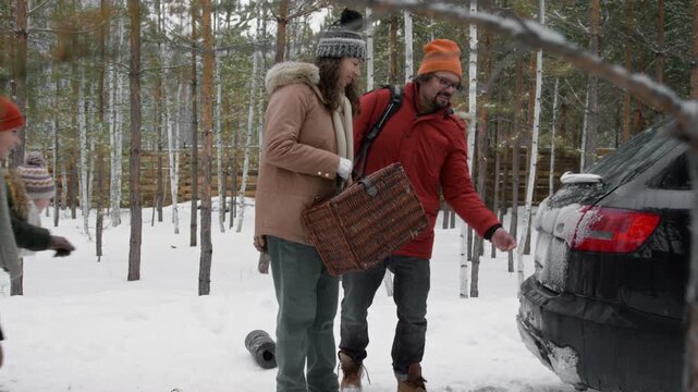 Full tracking shot of young Caucasian husband and wife carrying bags towards car, loading luggage into trunk, children helping, leaving to go home after spending vacation in winter forest cabin