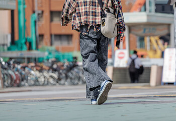Fashionably dressed in distressed plaid shirt and flared jeans, youth energetically struts across open plaza with crossbody bag on back and smartphone in hand.