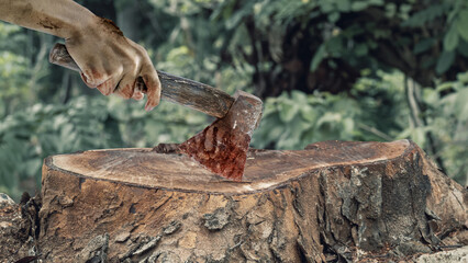 Zombie hand with wounds and blood holding an axe on a tree stump surface with blurred forest...