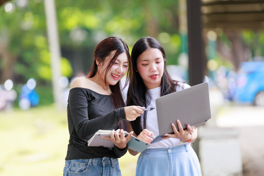 Two young Asian women students discussing project ideas on a laptop and notebook, collaborating outdoors on campus - Powered by Adobe