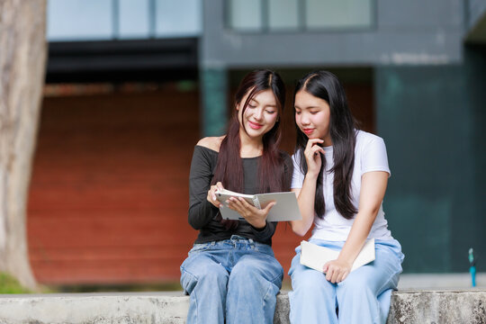 Two young Asian university friends collaborating on an assignment, reading a notebook and discussing while sitting outdoors on campus