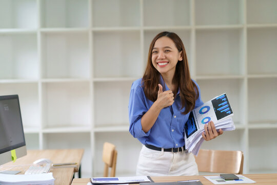 Smiling Asian businesswoman giving a thumbs up gesture, holding business charts and financial reports in a modern office