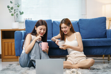Happy women friends relaxing, watching content on a laptop, and drinking coffee in casual clothes in the living room