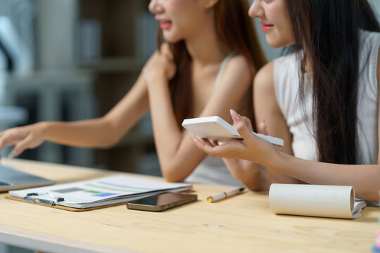 Two young Asian women concentrating, analyzing financial data and statistics together at an office desk, using a calculator and laptop