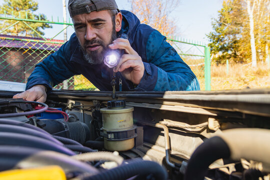 Man inspecting car engine with flashlight in sunny outdoor setting