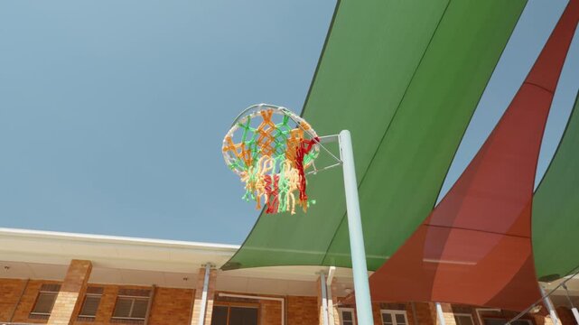 A ball is thrown and gets in the colourful basketball hoop with green and red shade sails in the background on a sunny blue sky day.