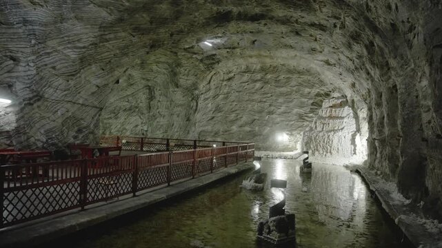 A static shot inside the massive Targu Ocna salt mine. A large cavern features rough-hewn salt walls, a wooden railing, and a pool of water reflecting the vaulted ceiling.