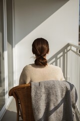 Peaceful morning scene of a woman sitting on a balcony in sunlight, wrapped in a cozy sweater and blanket, enjoying quiet solitude and minimalist comfort in a serene contemporary home setting