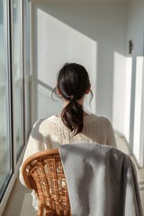 Peaceful morning scene of a woman sitting on a balcony in sunlight, wrapped in a cozy sweater and blanket, enjoying quiet solitude and minimalist comfort in a serene contemporary home setting