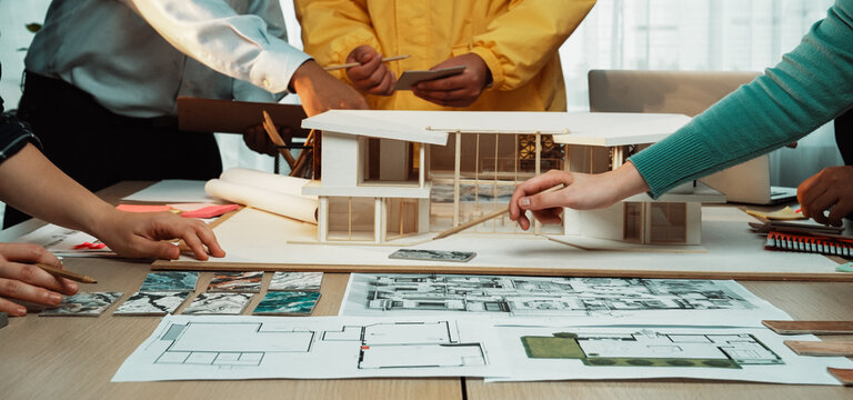 A diverse group of professionals collaboratively creating an architectural model in a modern office setting, surrounded by detailed blueprints and design plans. SACTR