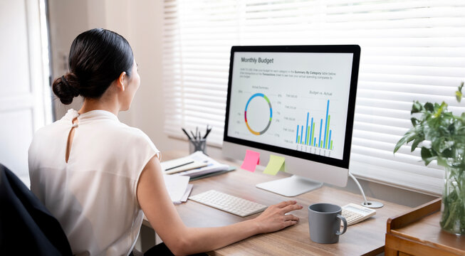 Young Asian businesswoman viewing a monthly budget report and financial charts on a desktop computer monitor in a modern office.