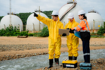 Team of environmental scientists or engineers in hazmat suits and PPE collecting water samples from a river for pollution analysis at an industrial site or factory.