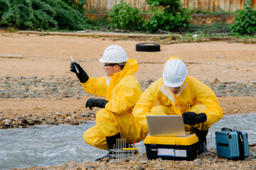 Team of environmental scientists or engineers in hazmat suits and PPE collecting water samples from a river for pollution analysis at an industrial site or factory.