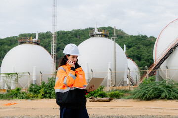 A woman wearing a hard hat and orange jacket is talking on her cell phone. Female engineer or supervisor in PPE at an industrial site (petrochemical plant, refinery, or shipping port)