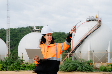 A woman wearing a hard hat and orange jacket is holding a laptop. Female engineer or supervisor in PPE at an industrial site (petrochemical plant, refinery, or shipping port)