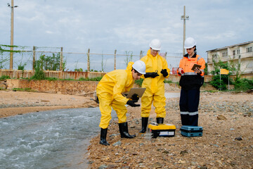 Team of environmental scientists in hazmat suits and a female engineer/supervisor collaborating on water pollution analysis and inspection at an industrial plant site.