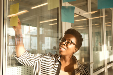 Obraz premium Black woman in glasses standing in modern office, looking at sticky notes stuck on glass wall and smiling cheerfully