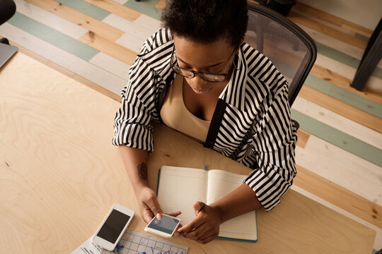 Stylish African American woman in striped blouse checking social media on smartphone while sitting at table in modern office - Powered by Adobe