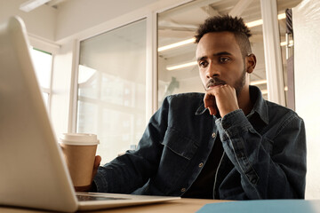 Portrait of young African American man in denim shirt reading news on laptop while sitting at table with coffee in modern office, low angle view