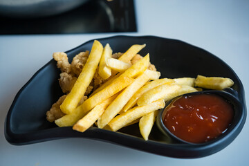 French fries with ketchup served on a black plate alongside crispy fried chicken pieces on a white table surface
