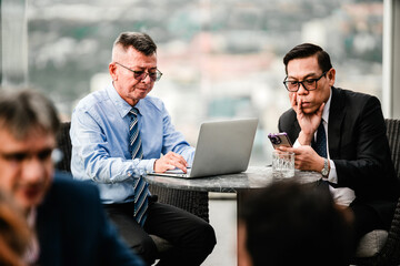 Two business professionals in formal attire work at a cafe lounge table, one on a laptop and the other on a smartphone, in an urban office setting during a candid collaborative meeting.