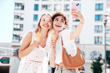 Two young beautiful smiling hipster female in trendy summer clothes. Carefree women posing on street background. Positive models eating tasty ice cream in waffles cone in sunny day, take selfie photo