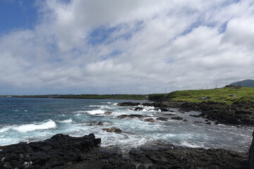 Waves crash over black basalt rocks along grassy coastline under broken clouds, Rishiri Island, Hokkaido, Japan