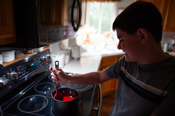 Teen boy carefully stirs red candy mixture on home kitchen stove