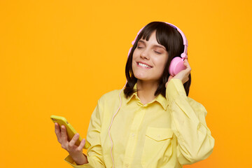 A young woman in a pale yellow shirt wears pink headphones and smiles with eyes closed while...