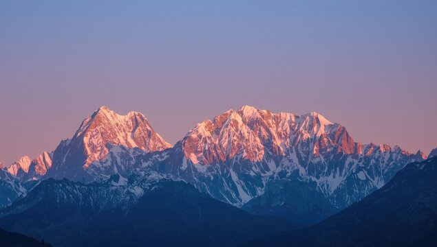 Majestic snow-capped mountain range basks in the warm glow of sunrise or sunset, with stunning hues of pink and orange illuminating the peaks against a soft, pastel blue sky.