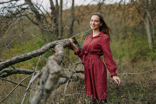 Young woman in a red dress stands by a fallen tree in a natural meadow, captured in an authentic moment that emphasizes genuine expression, credible portrayal and relaxed posture.