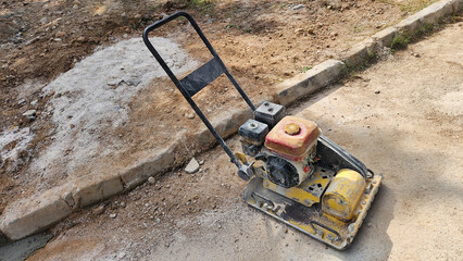Plate compactor rests on  gravel next to a curb, used for construction or paving work.