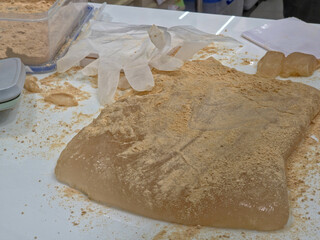 A large, translucent piece of gelatinous mochi or Warabi Mochi dusted heavily with Kinako (toasted soybean flour) on a messy preparation counter with plastic gloves.