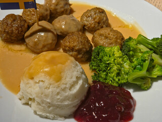 Close-up of a plate of classic Swedish meatballs (Köttbullar) served with cream sauce, mashed potatoes, lingonberry jam, and steamed broccoli, topped with a Swedish flag pick.