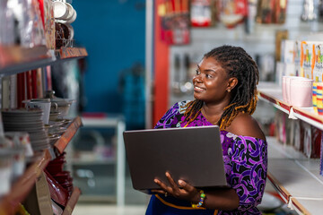 African businesswoman, SME owner in her vibrant supermarket aisle, reviewing inventory with her laptop computer