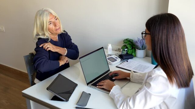 Senior woman at a medical appointment explaining her symptoms and pointing to her painful arm while the female doctor listens attentively and takes notes on her laptop in the clinic office