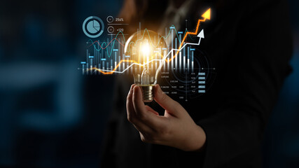 A businesswoman holds a glowing light bulb, symbolizing innovation, amidst digital financial...